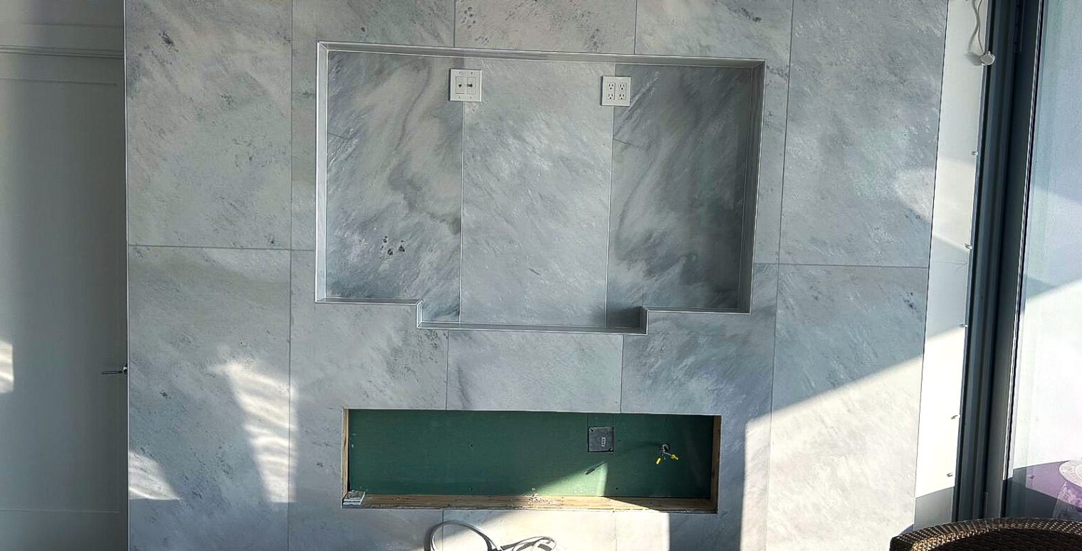 A marble wall installation elegantly frames two inset spaces for a TV and speakers, ideal for any remodeling project. Featuring convenient electrical outlets, the design includes a black shelf below and a lower recessed area. Sunlight casts shadows across the scene with a bundle of cables and lamp on the shelf.