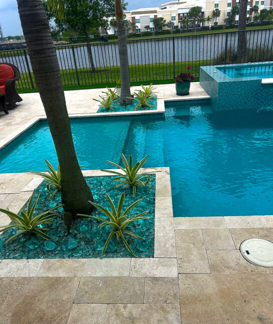 A backyard pool with clear blue water, bordered by palm trees and decorative plants. The area showcases light-colored tiled flooring. A wrought iron fence offers a view of a lake and buildings in the background, all beneath a cloudy sky, perfect for an elegant remodeling inspiration.