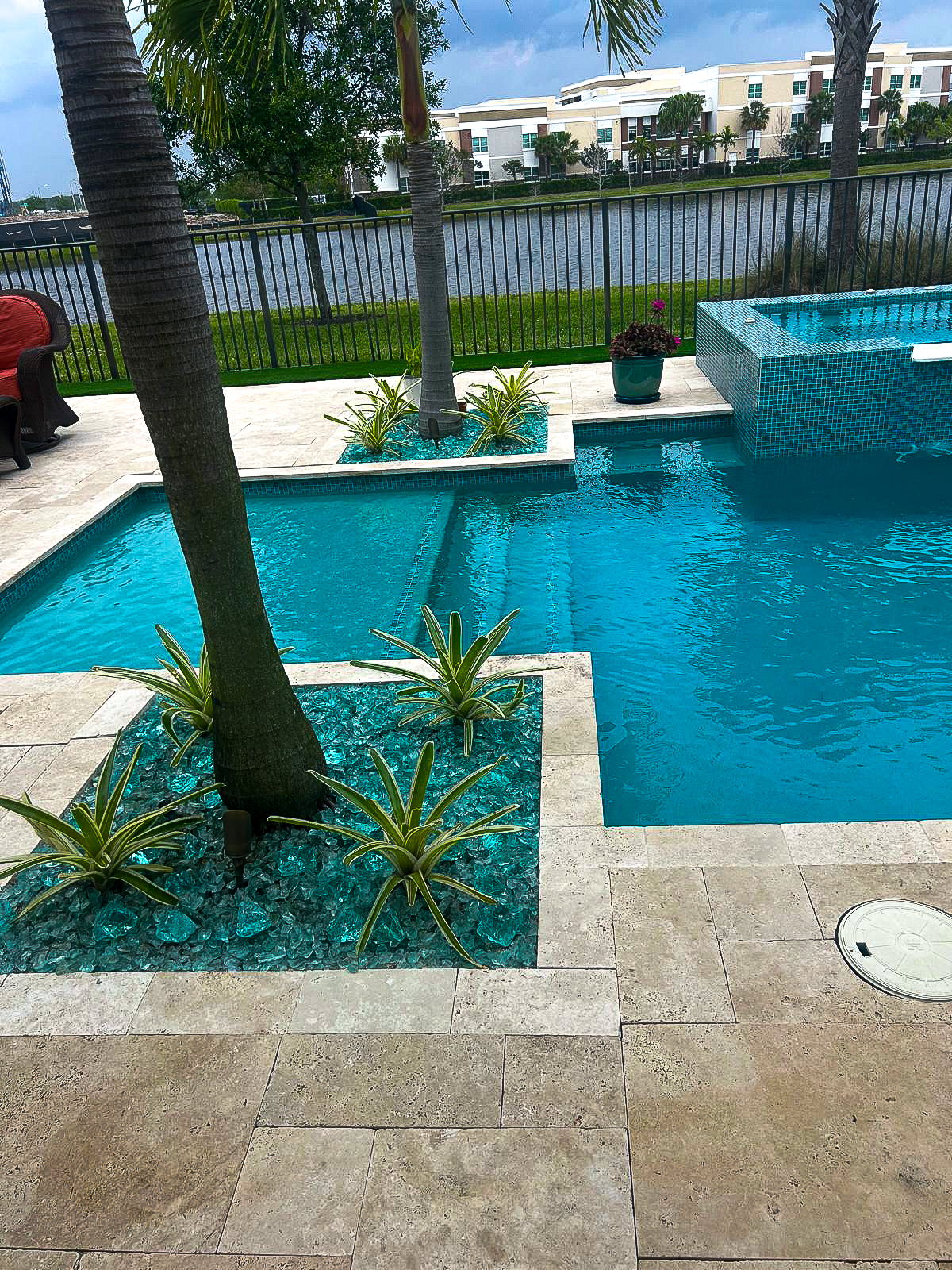 A backyard pool with clear blue water, bordered by palm trees and decorative plants. The area showcases light-colored tiled flooring. A wrought iron fence offers a view of a lake and buildings in the background, all beneath a cloudy sky, perfect for an elegant remodeling inspiration.