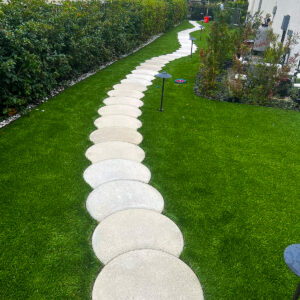 A winding path of large circular stepping stones elegantly curves through a backyard with green grass, a testament to thoughtful renovation. The path is bordered by hedges on one side and a garden area on the other, with houses and a small lamp visible in the background.