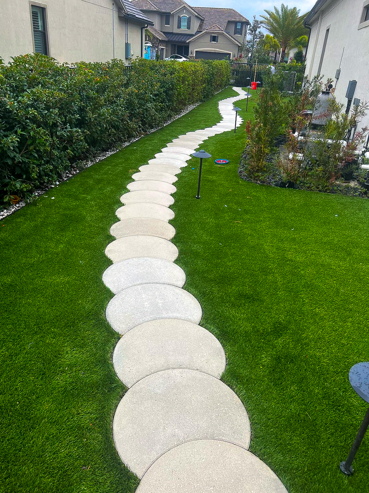 A winding path of large circular stepping stones elegantly curves through a backyard with green grass, a testament to thoughtful renovation. The path is bordered by hedges on one side and a garden area on the other, with houses and a small lamp visible in the background.