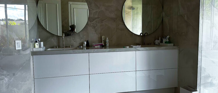 A modern bathroom with two round mirrors above a white double-sink vanity showcases expert renovation. Various toiletries are on the counter, and the tiled walls complement the stylish flooring. Two white bath mats adorn the floor while a window reflects natural light.