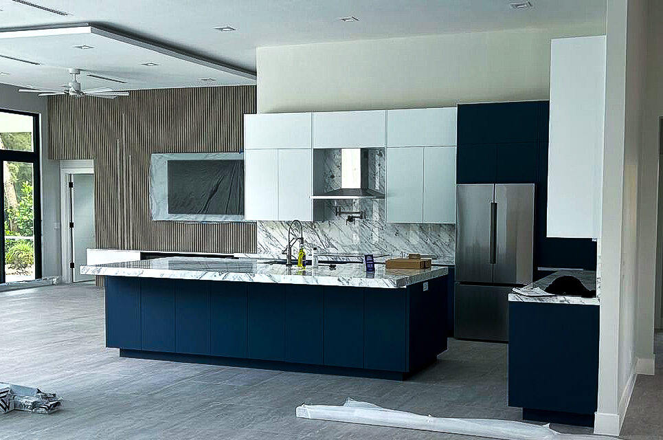 Modern kitchen with dark blue and white cabinetry. A large marble island counter sits centrally. Stainless steel fridge and a marble backsplash accentuate the space. Floor-to-ceiling windows allow natural light to fill the area.