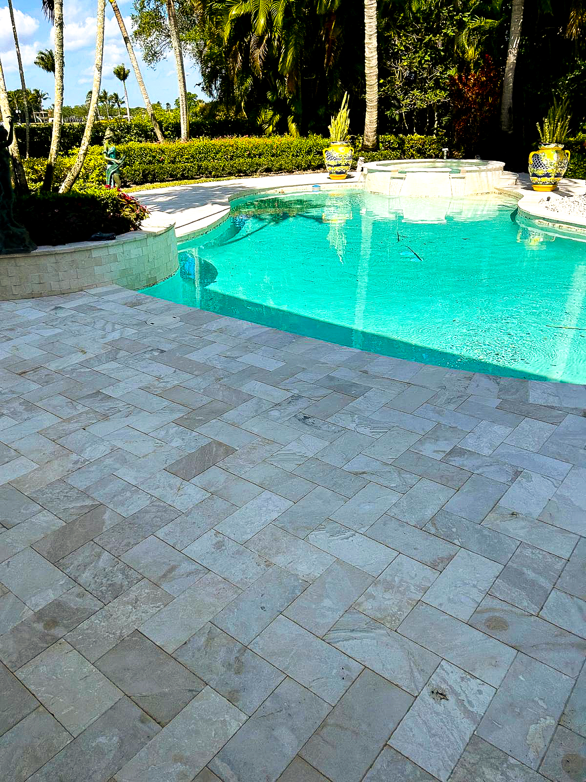 A luxurious outdoor pool with a light stone deck is surrounded by palm trees and lush greenery, creating an oasis that makes you forget you ever considered remodeling. Bright blue water shimmers under a clear sky, flanked by two decorative urns with plants.