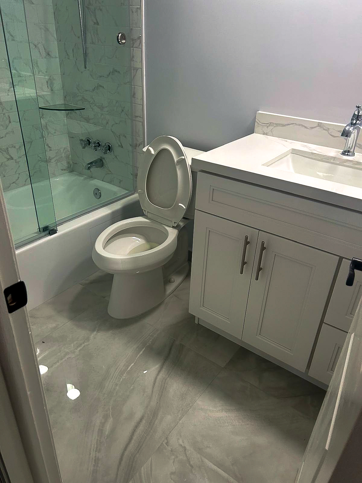 A bathroom with a marble-patterned floor features a white toilet with its lid open. Next to it is a white vanity with a sink and chrome faucet. The bathtub has a glass shower screen and marble-style tiles.