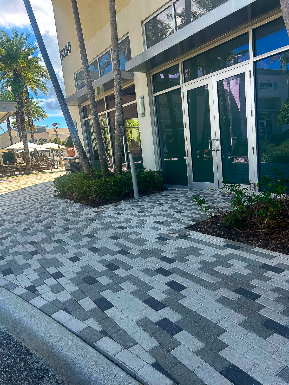 A modern building with large glass doors stands next to a paved pedestrian walkway featuring a sleek gray and white geometric flooring pattern. Palm trees and greenery line the path. In the background, outdoor seating is visible under umbrellas, suggesting recent renovation efforts.