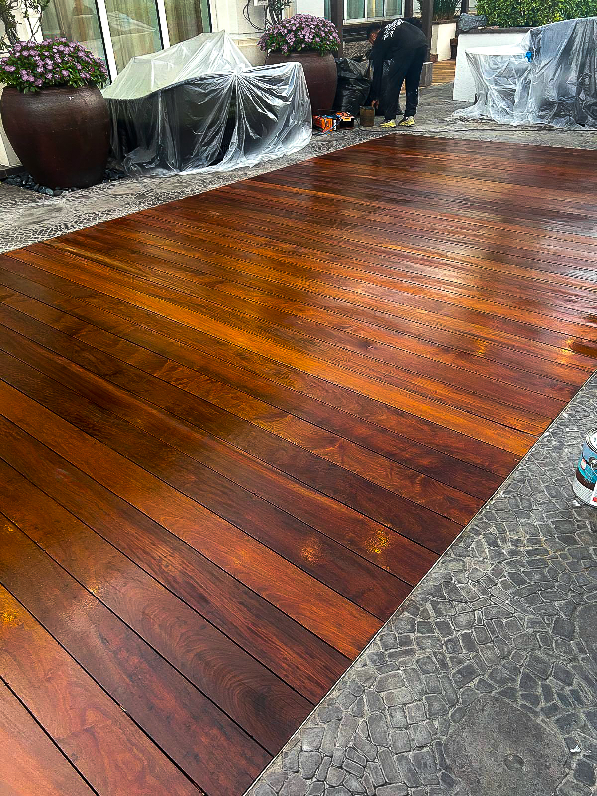 A freshly polished wooden deck glistens in the sunlight, hinting at a recent renovation. Large potted plants line the background, and furniture is covered with protective sheets. A person is working in the background, partially visible near the edge.