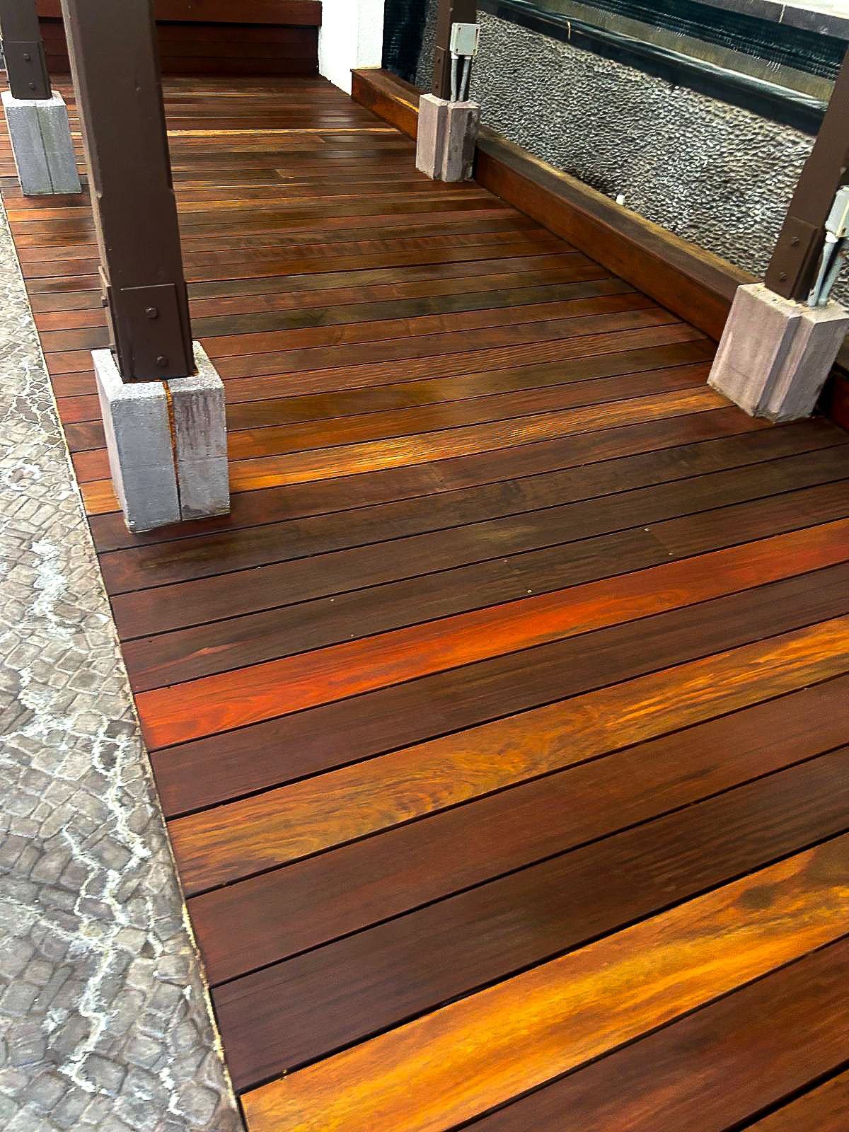 A wooden deck with rich brown and reddish planks complements a textured stone tile walkway, showcasing the art of modern outdoor flooring. Vertical beams supported by concrete bases add to this inviting renovation, blending materials for a seamless remodeling of your outdoor space.