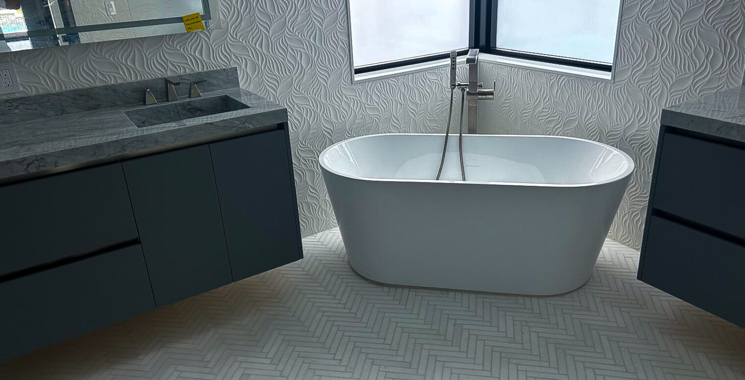 Modern bathroom featuring a white freestanding tub near a corner window, surrounded by textured walls. Dual gray vanities with marble countertops are on either side. The herringbone-patterned floor complements the elegant, minimalist design.