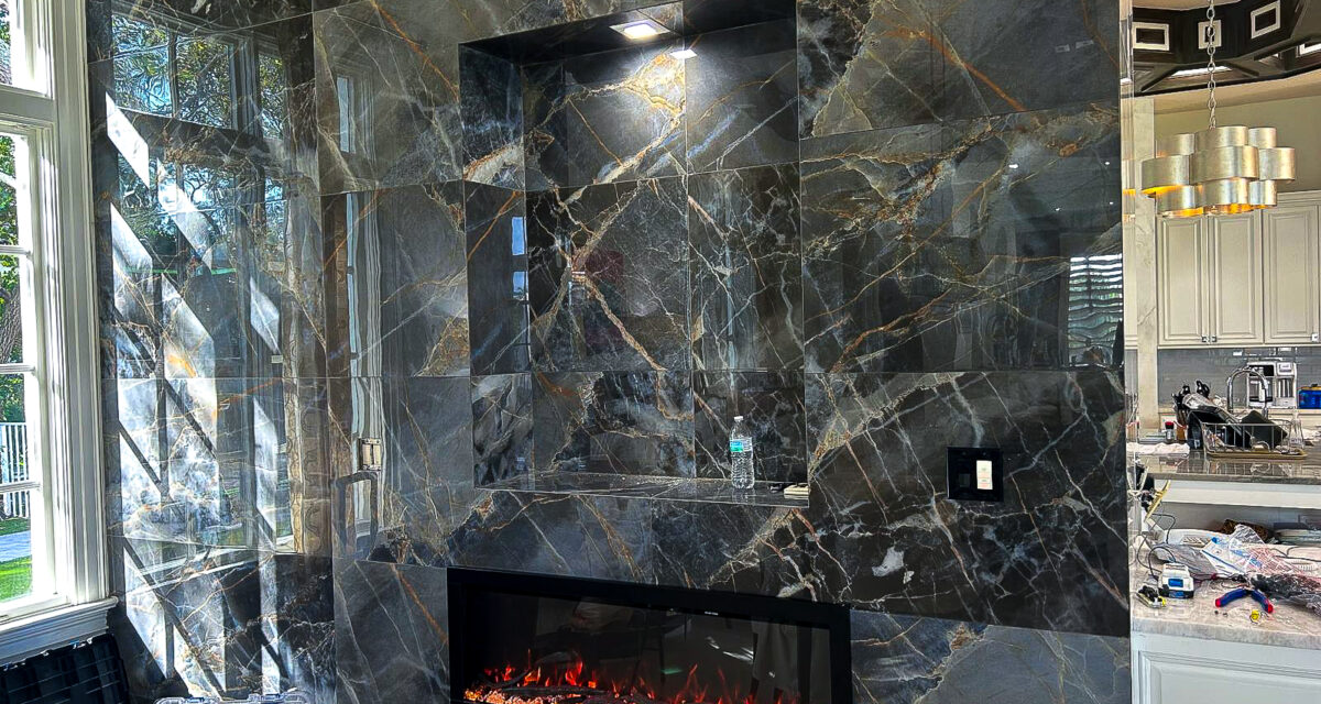 Luxurious living room with a dark marble accent wall featuring a built-in electric fireplace. The marble has striking white and gold veining. To the right, part of a modern kitchen is visible with sleek cabinets and elegant lighting.