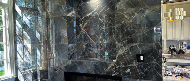 Luxurious living room with a dark marble accent wall featuring a built-in electric fireplace. The marble has striking white and gold veining. To the right, part of a modern kitchen is visible with sleek cabinets and elegant lighting.