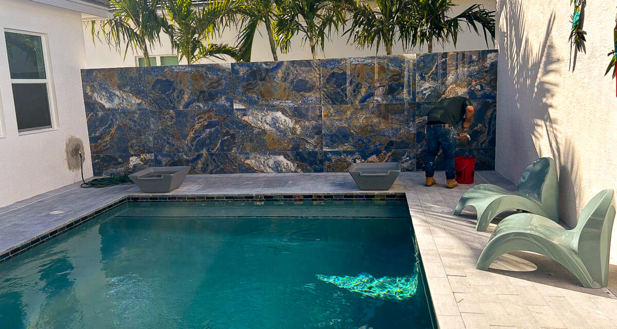 A backyard with a rectangular pool, surrounded by stone tiles. Two modern lounge chairs are on the right. A person is tending to items near a large decorative stone wall. Palm trees are visible in the background.