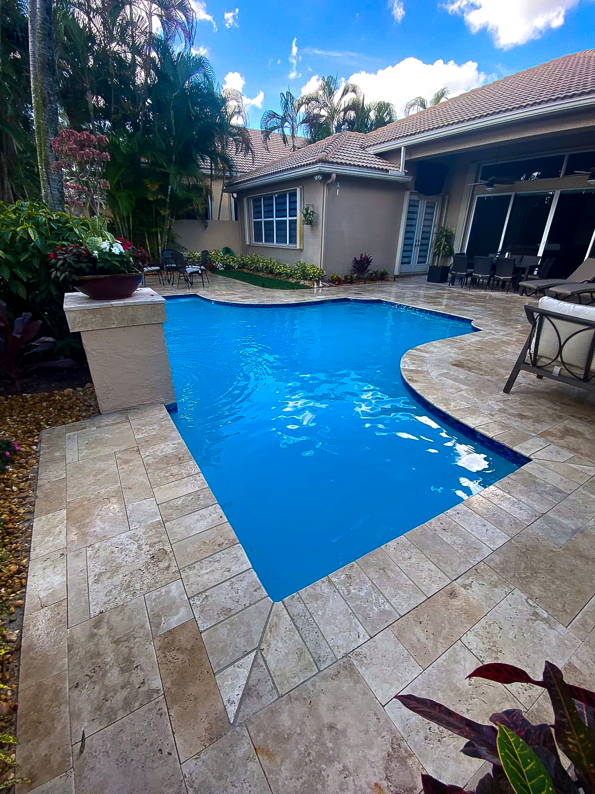 A backyard with a small, curved swimming pool surrounded by beige stone tiles. The area is adorned with lush greenery and palm trees. Lounge chairs and a table are set up on the patio near the pool. The sky is partly cloudy.