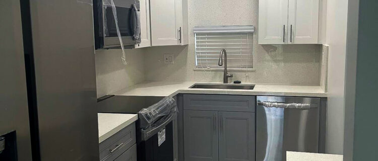 Modern kitchen with light gray and white cabinets, stainless steel appliances, and beige countertops. Features include an oven, microwave, dishwasher, and refrigerator. Recessed lighting illuminates the space, and theres a small window above the sink.