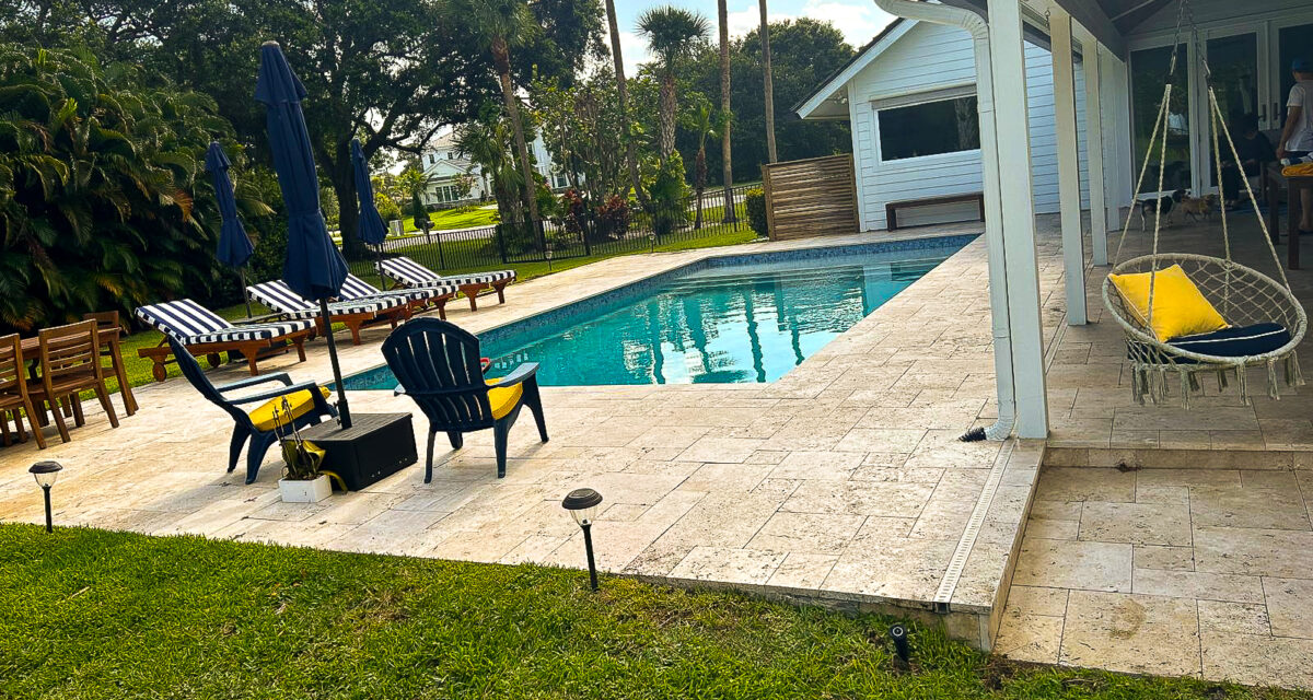 A backyard scene featuring a swimming pool with a stone deck surrounded by greenery. There are lounge chairs with striped cushions, a table with a yellow towel, and a hammock chair. A white house stands nearby under a partly cloudy sky.