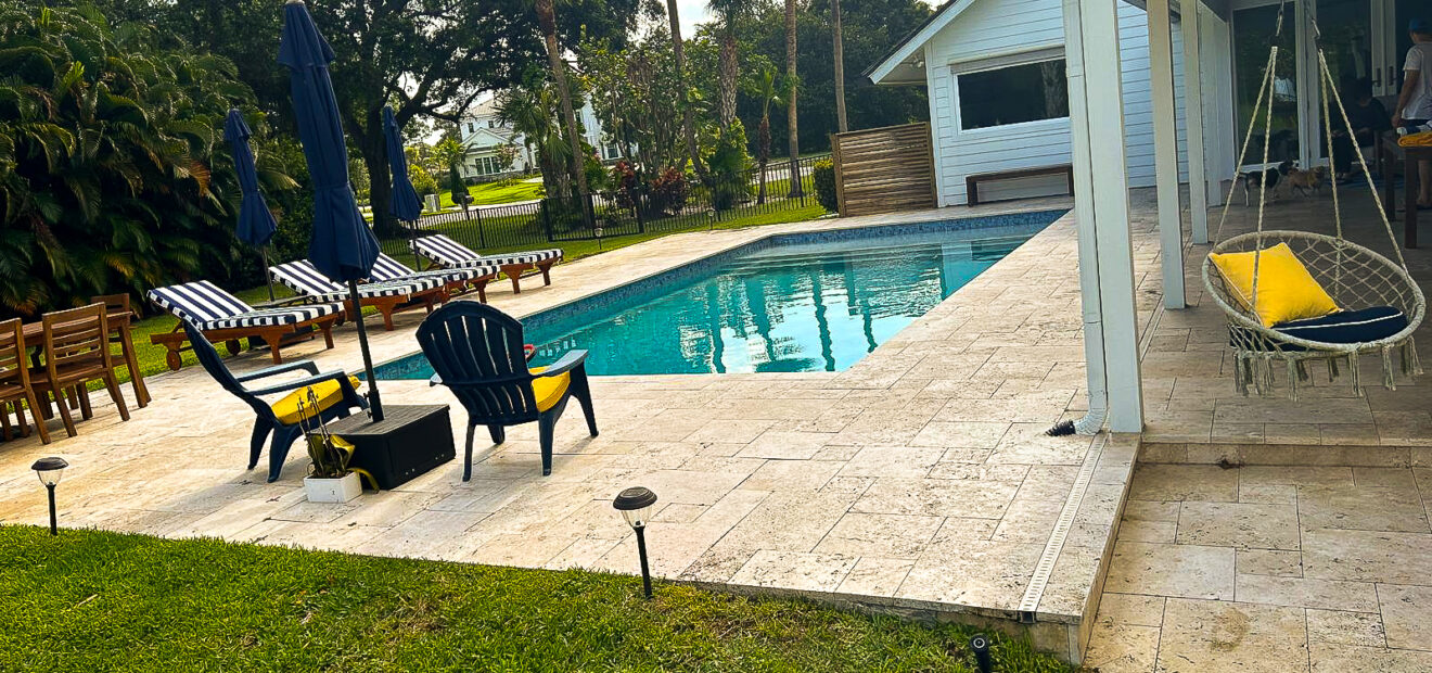 A backyard scene featuring a swimming pool with a stone deck surrounded by greenery. There are lounge chairs with striped cushions, a table with a yellow towel, and a hammock chair. A white house stands nearby under a partly cloudy sky.