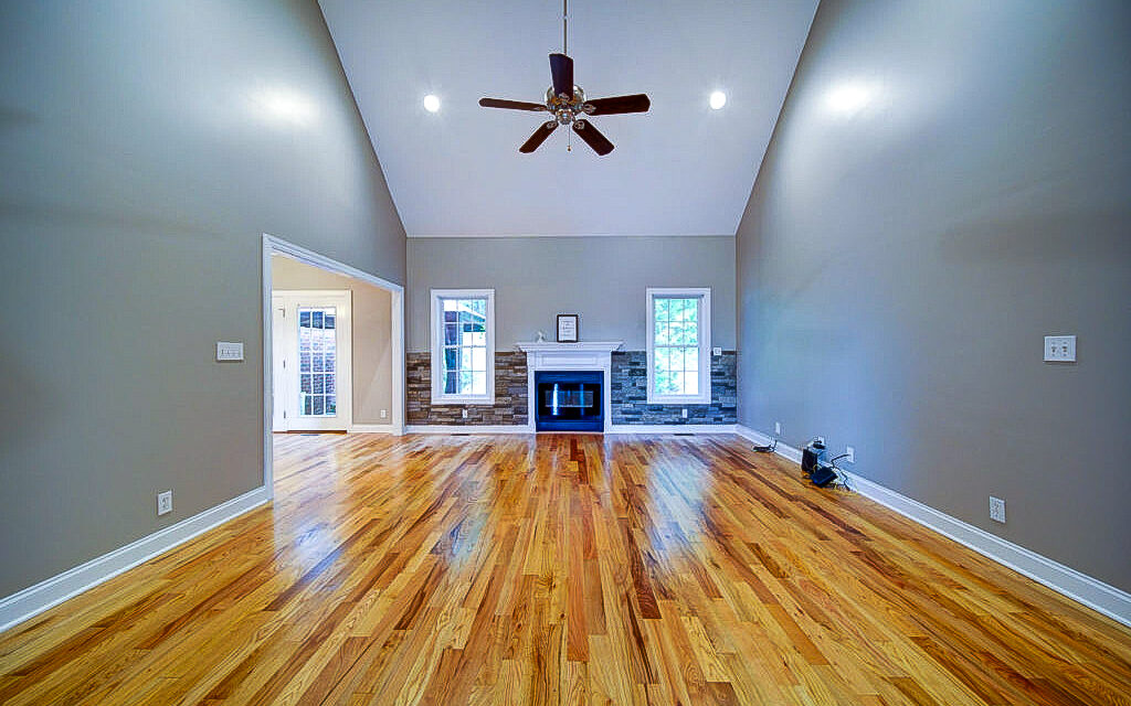 Spacious living room with hardwood floors, a vaulted ceiling, and a central ceiling fan. A fireplace is centered between two windows, and light gray walls enhance the airy atmosphere. An open doorway leads to another room.