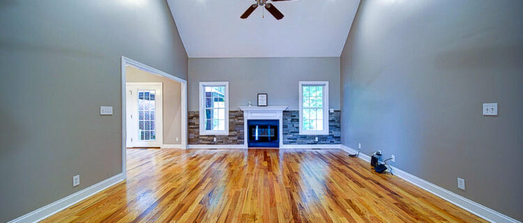 Spacious living room with hardwood floors, a vaulted ceiling, and a central ceiling fan. A fireplace is centered between two windows, and light gray walls enhance the airy atmosphere. An open doorway leads to another room.