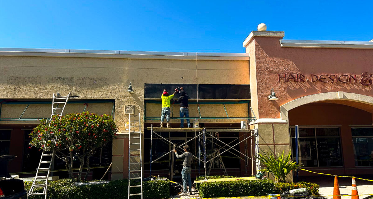 Workers on scaffolding install signage on a storefront. One worker is on top, while another guides from below. Ladders and safety cones are visible. The sky is clear, and theres a tree and plants near the entrance.