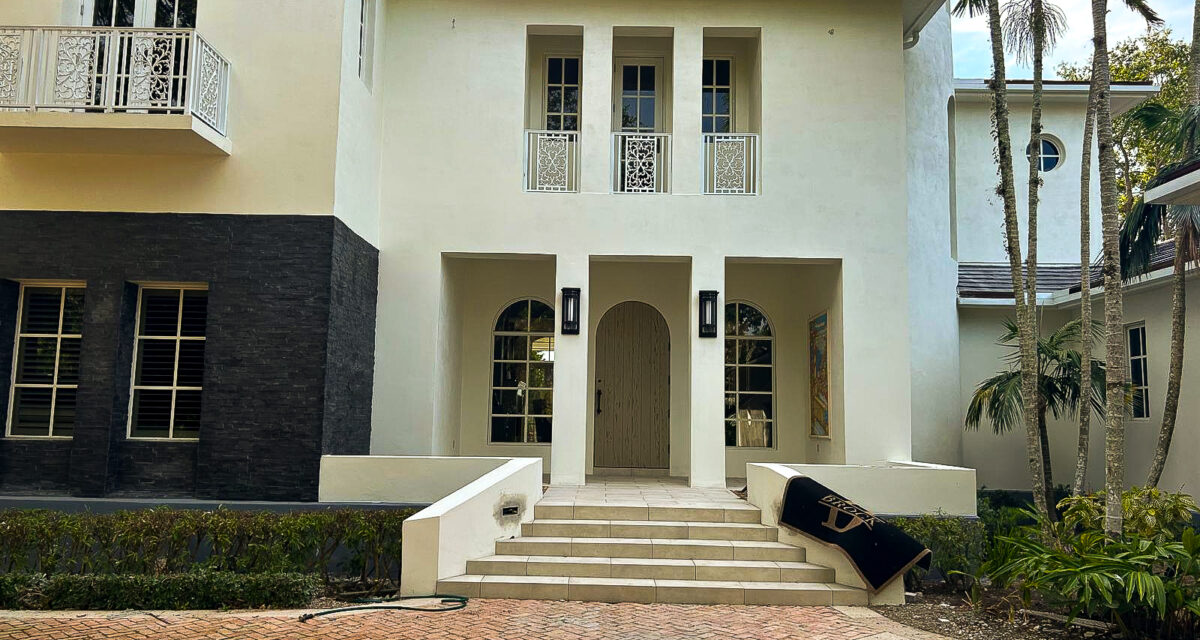 A large, modern white house with an arched wooden front door, surrounded by tall windows and black wall accents. A staircase with low white walls leads to the entrance. Tropical trees frame the house, and a brick driveway is in the foreground.