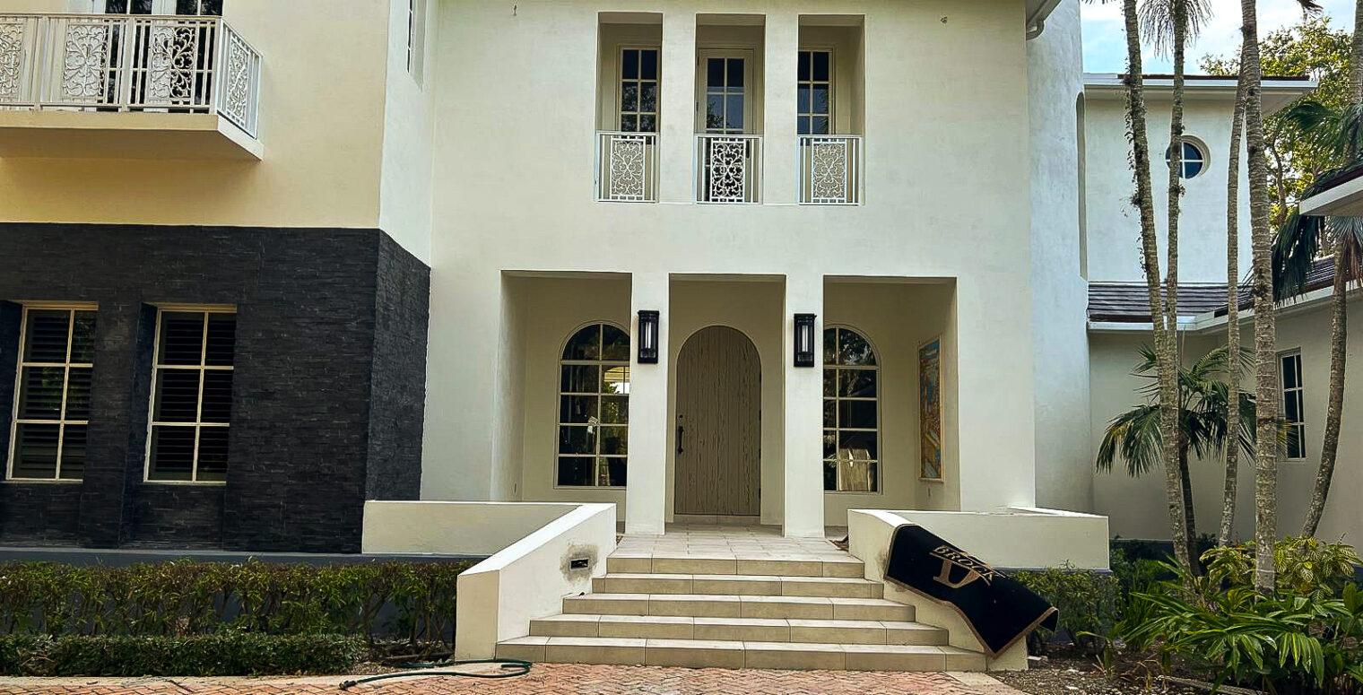 A large, modern white house with an arched wooden front door, surrounded by tall windows and black wall accents. A staircase with low white walls leads to the entrance. Tropical trees frame the house, and a brick driveway is in the foreground.