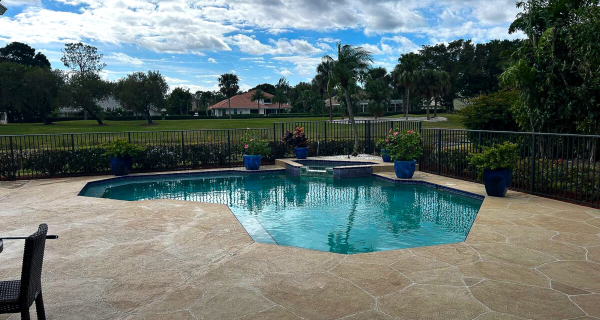 A backyard pool with hexagonal stone tiles surrounds the water. Blue pots with plants line the edge. The pool area is enclosed by a black fence. In the background, palm trees and houses can be seen under a partly cloudy sky.