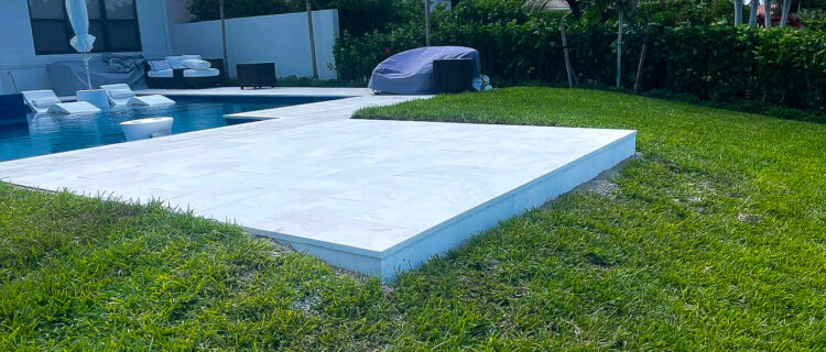 A backyard with a swimming pool surrounded by a white patio. There are lounge chairs and an umbrella nearby. Green grass borders the patio, and palm trees and houses are visible in the background under a blue sky.