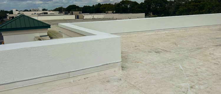 Rooftop with a flat white surface, surrounded by short white walls. In the distance, there are trees and buildings under a partly cloudy sky. A small vent or pipe is visible on the roof.