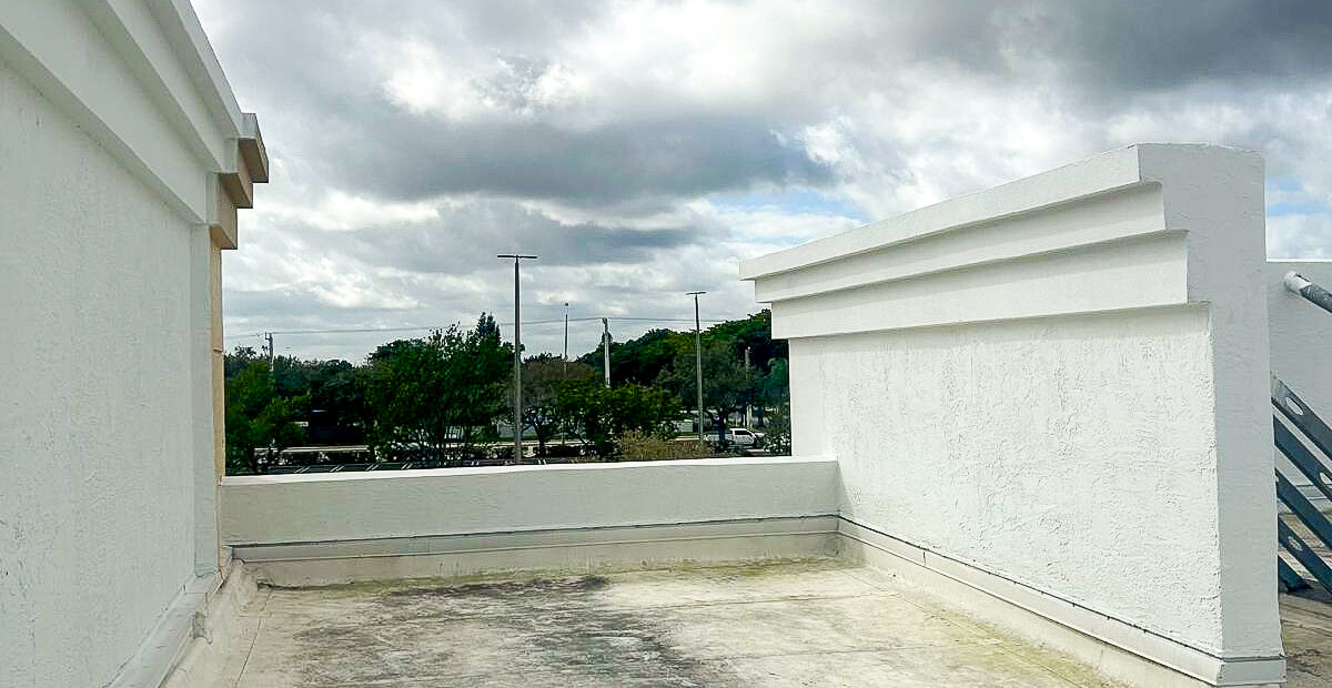 A deserted rooftop with white walls under a cloudy sky. The ground is slightly dusty, and there are some trees and a parking lot visible in the background.