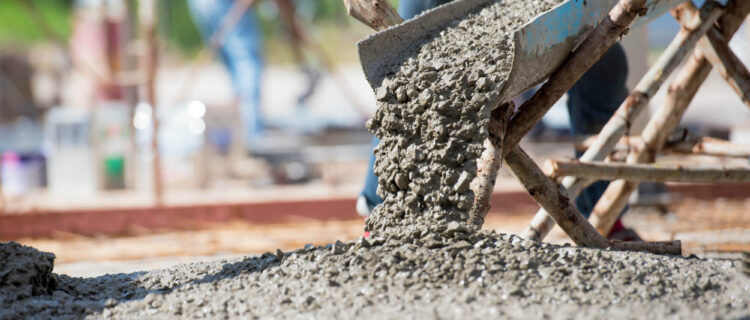 Concrete being poured from a chute at a construction site, with a worker visible in the background. The mixture spreads on the ground, surrounded by wooden scaffolding and blurred greenery.