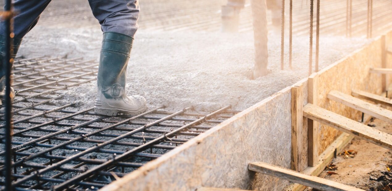 A construction worker in boots pours concrete onto a metal rebar grid for a foundation. The sun casts a warm glow, and wooden supports are visible on the sides.