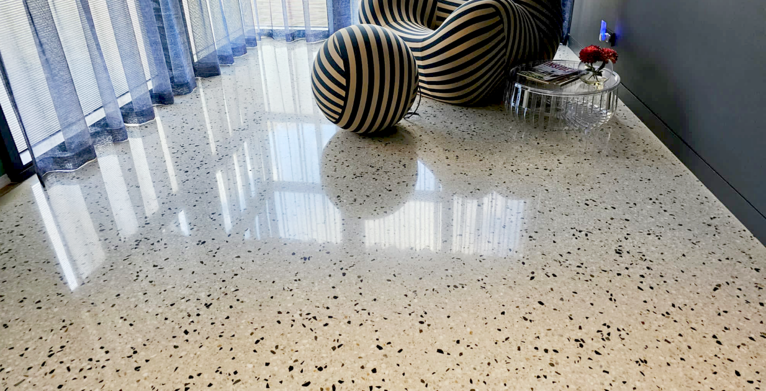 A polished terrazzo floor with a smooth, speckled pattern is shown. A modern striped chair and small round glass table with a plant are in the corner near large windows with sheer curtains, allowing natural light to illuminate the space.