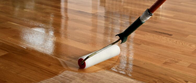 A paint roller applies a glossy finish to a wooden floor, enhancing the wood grains shine and texture. The roller has a red end and a handle with a black grip, reflecting light from the polished surface.